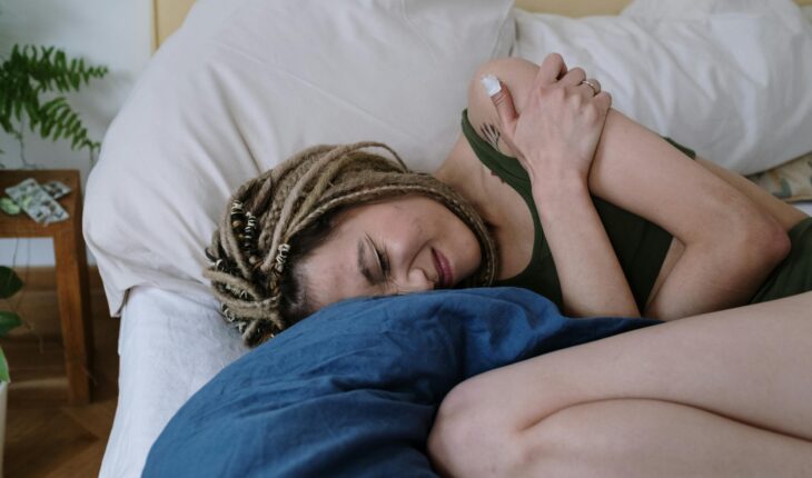 A woman with dreadlocks lying in bed holding her stomach in pain, possibly due to cramps.