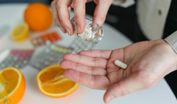 Close-up of a person taking a vitamin capsule with citrus fruit and medication on a table.